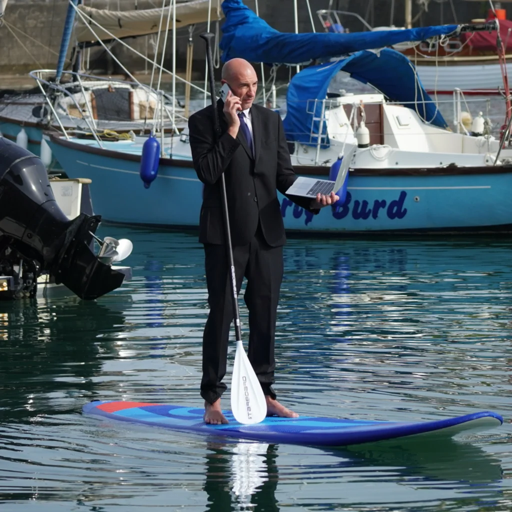 Barefoot businessman in suit stands on paddleboard in marina, holding paddle, phone to ear and open laptop.