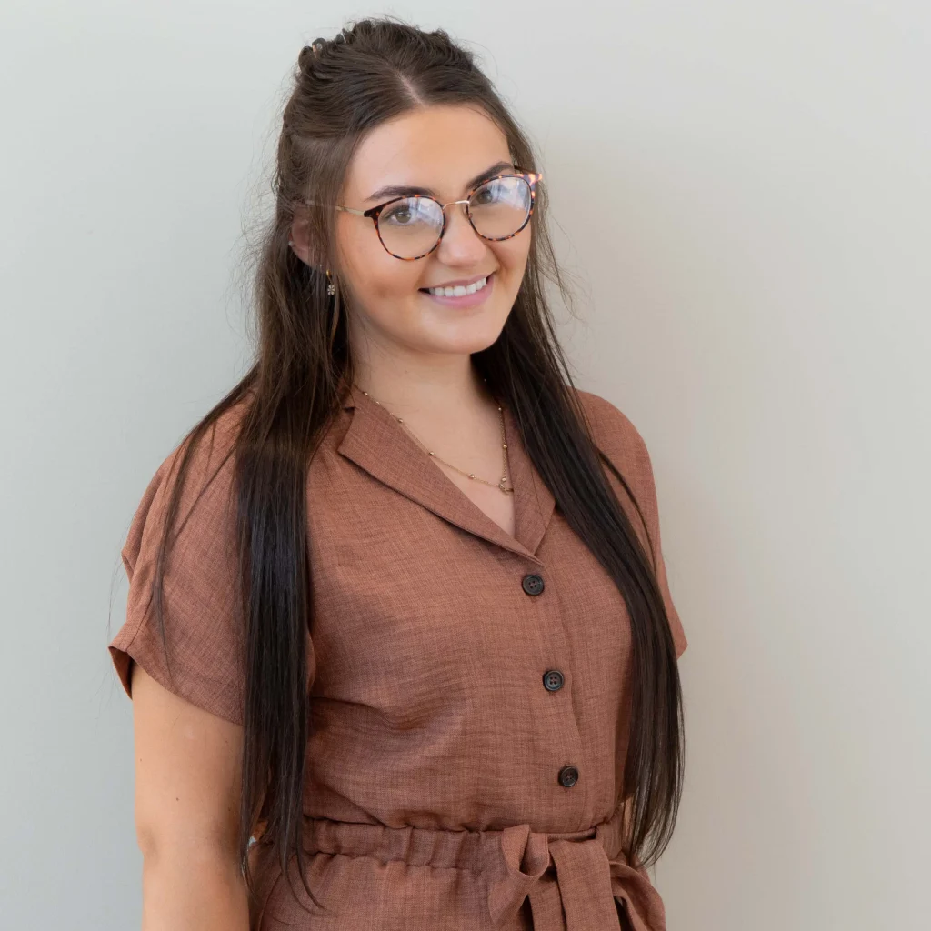 Smiling woman with long dark hair and glasses wearing brown belted button-up dress against light background.