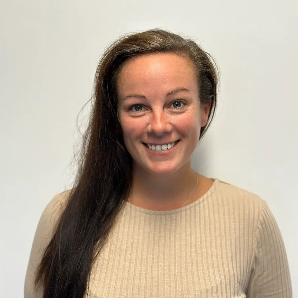 Smiling woman with long dark hair swept to the side wearing beige ribbed top against plain light background.