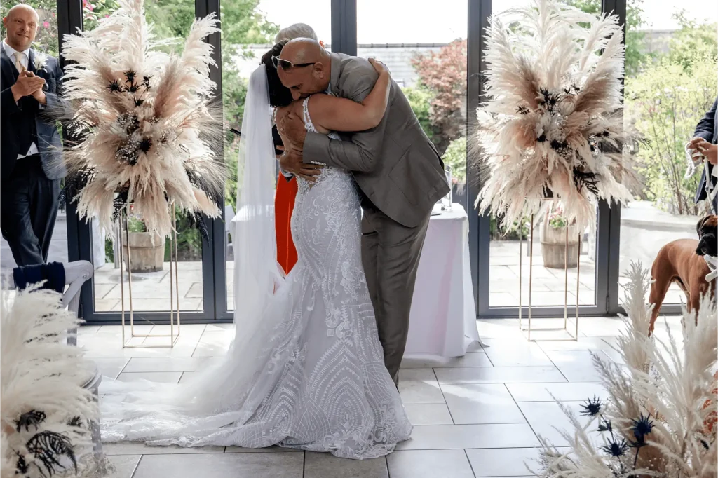 Bride and groom share an emotional hug during indoor wedding ceremony with pampas grass floral arrangements.