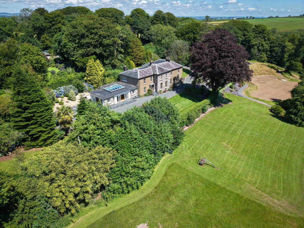 Aerial view of a Cilrhiw Country House with manicured lawns, gardens, and surrounding trees under a sunny sky.