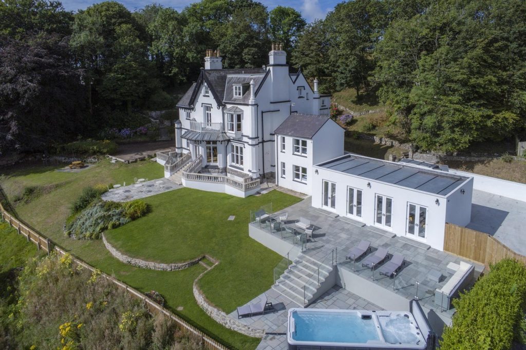 Large white countryside house with patio, hot tub, and manicured lawn, surrounded by trees and gardens.