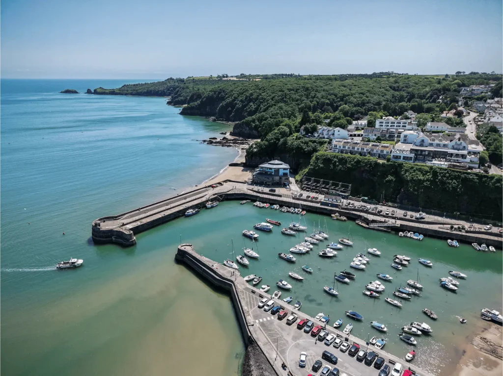 Aerial view of a coastal harbor with boats docked, a curved pier, and buildings along the cliffside under clear blue skies.