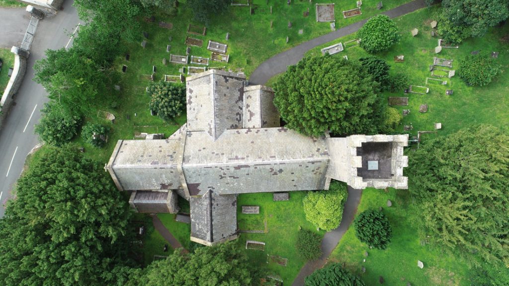 Aerial view of an old stone church with cross-shaped layout, surrounded by trees and gravestones in a green cemetery.