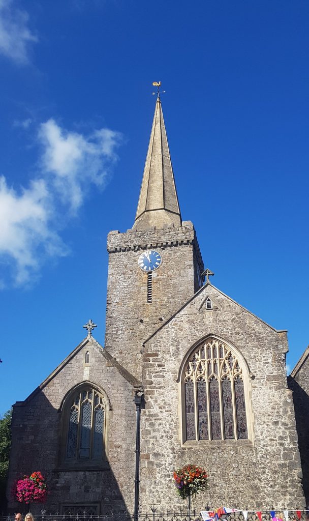 Stone church with tall steeple, clock face, stained glass windows, and hanging flower baskets under a clear blue sky.