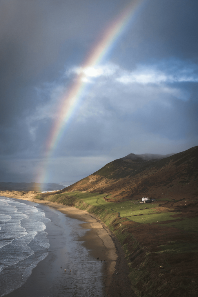 A vibrant rainbow arches over the sandy beach in Wales, creating a stunning coastal scene