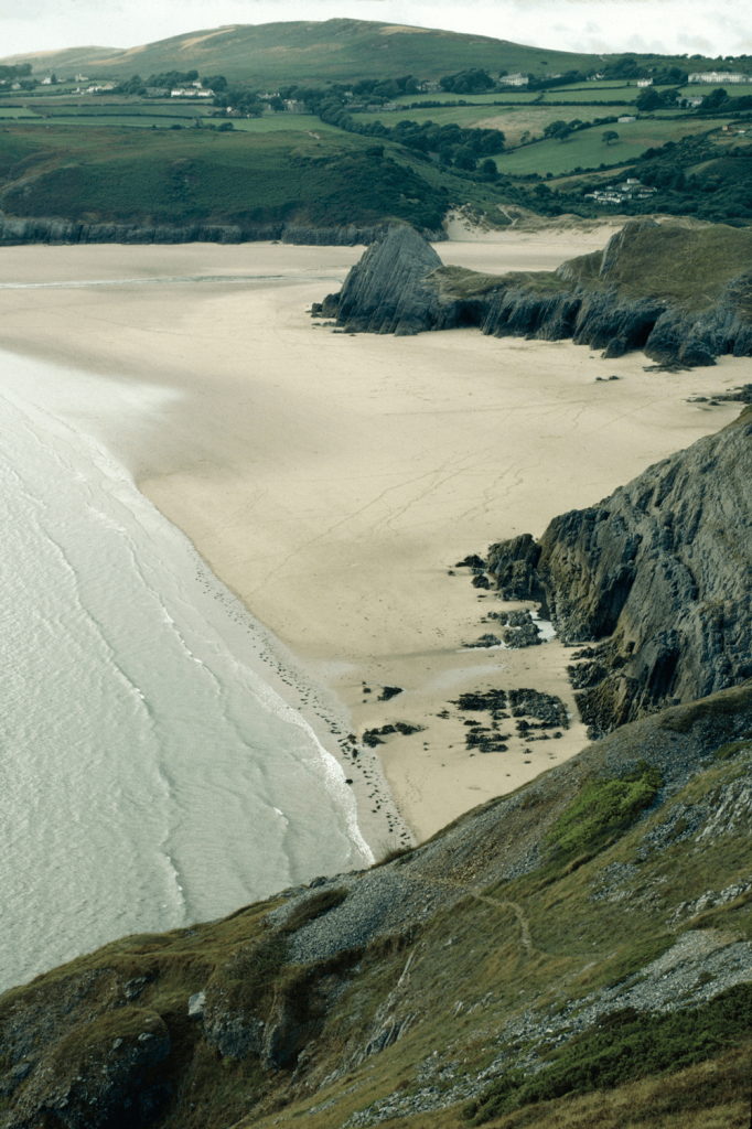 A beautiful sandy beach stretches out, framed by rolling grassy hills and a bright, sunny sky above.